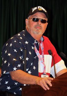 Dwight pictured at a podium wearing an American flag shirt and Air Force hat
