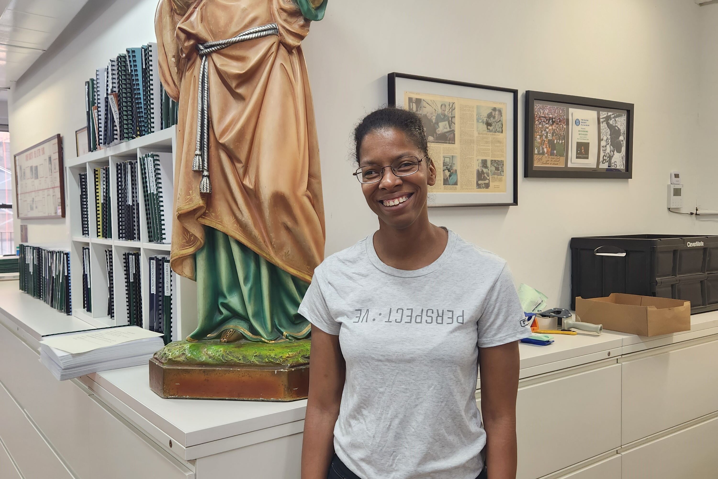 Tiffany, a young woman with glasses, pictured in front of our St. Lucy statue in our office