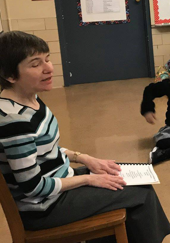 Mary seated reading a braille book to school children